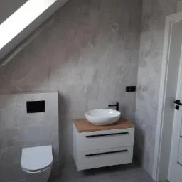 Nowoczesna łazienka na poddaszu z szarymi płytkami imitującymi marmur, białą wiszącą toaletą i umywalką nablatową na szafce z drewnianym blatem, czarna armatura, widoczne okablowanie na ścianie.