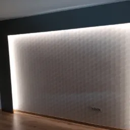 Ściana z trójwymiarową tapetą w geometryczne wzory, oświetlona od tyłu listwą LED, tworząca efektowną dekorację wnętrza. Ciemnoszara rama wokół podświetlonej ściany, drewniana podłoga.