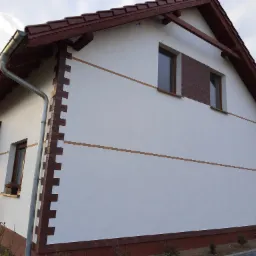 Elewacja domu jednorodzinnego z białym tynkiem, brązową cegłą klinkierową na narożnikach i cokole, oraz brązową dachówką i orynnowaniem, widok z boku.