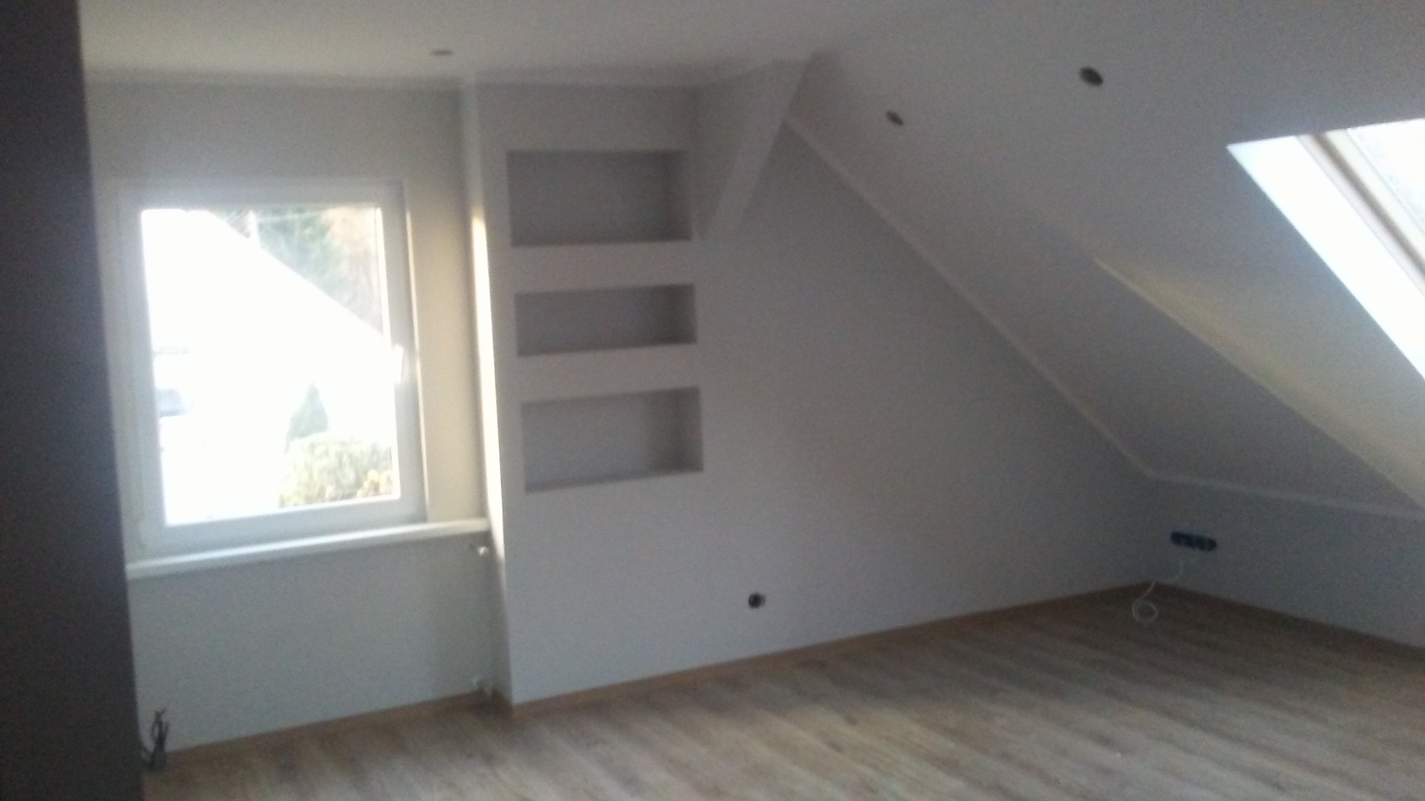 Poddasze po remoncie z oknem dachowym, drewnianą podłogą i wnękami w ścianie pomalowanymi na biało.