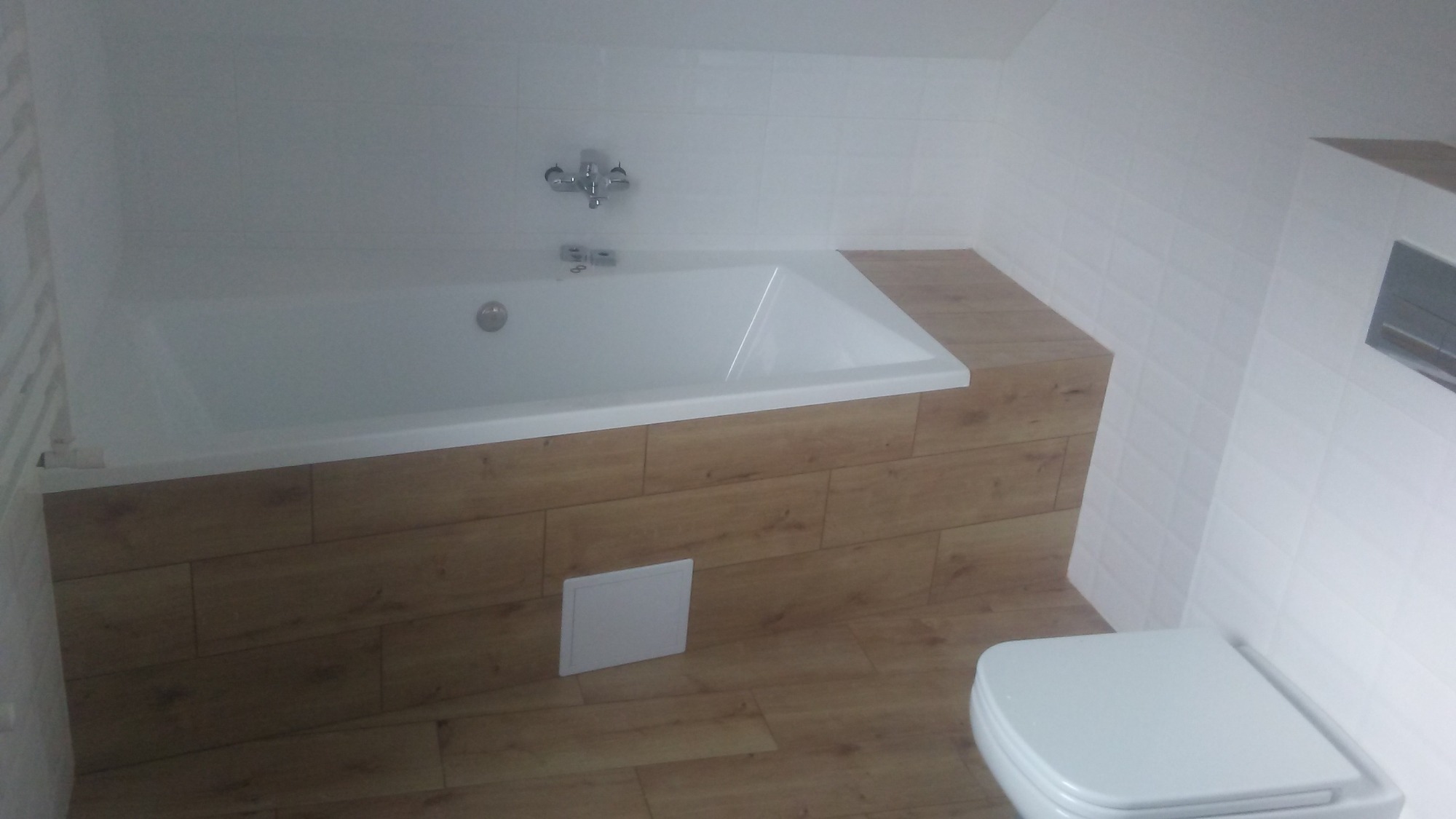 Biała wanna z chromowaną baterią, obudowana panelami imitującymi drewno, białe płytki na ścianach, toaleta z deską wolnoopadającą i podłoga wyłożona panelami w odcieniach drewna.