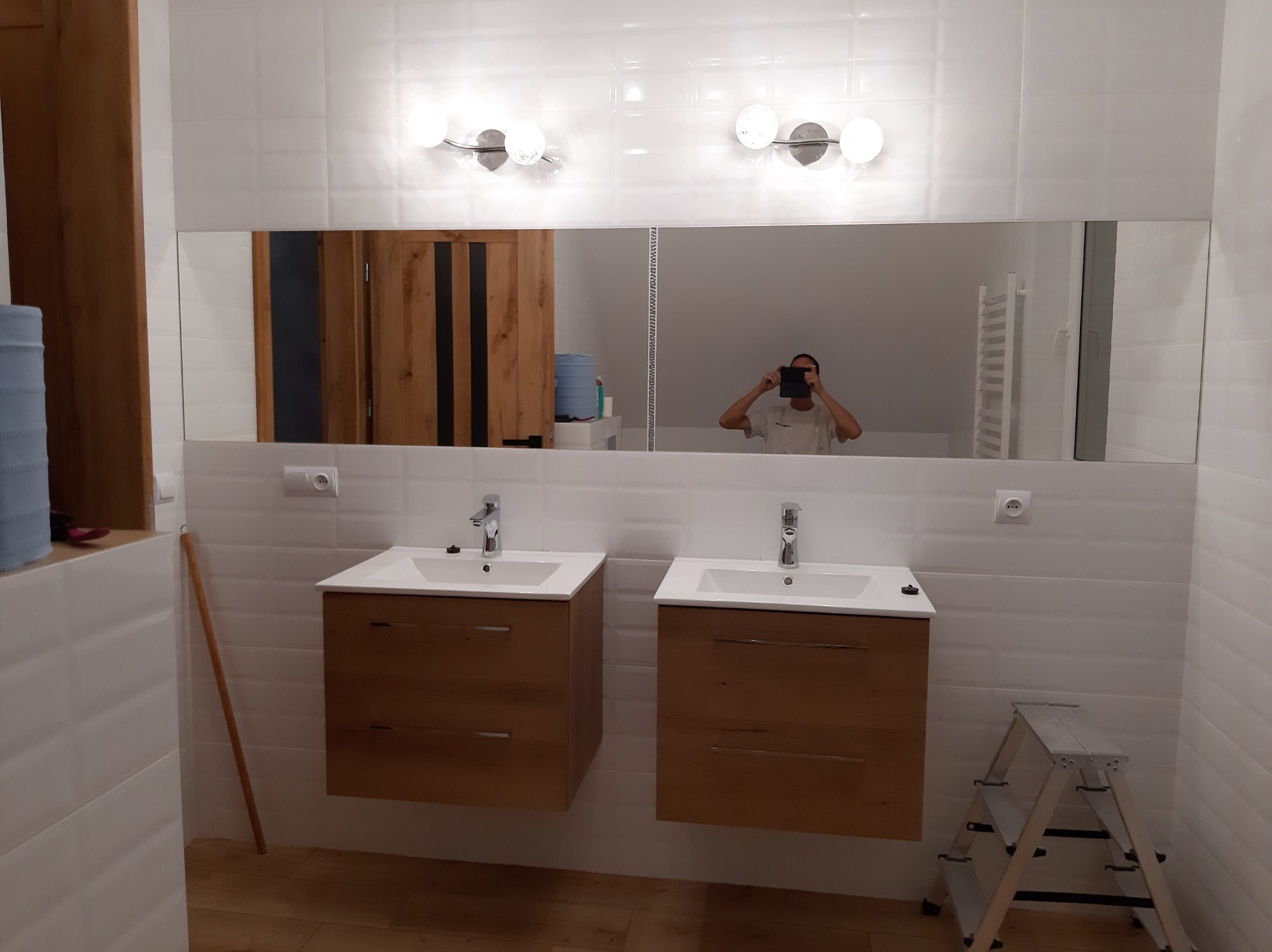 Nowoczesna łazienka z dwoma umywalkami na drewnianych szafkach, duże lustro z oświetleniem, białe płytki na ścianach i drabina w rogu