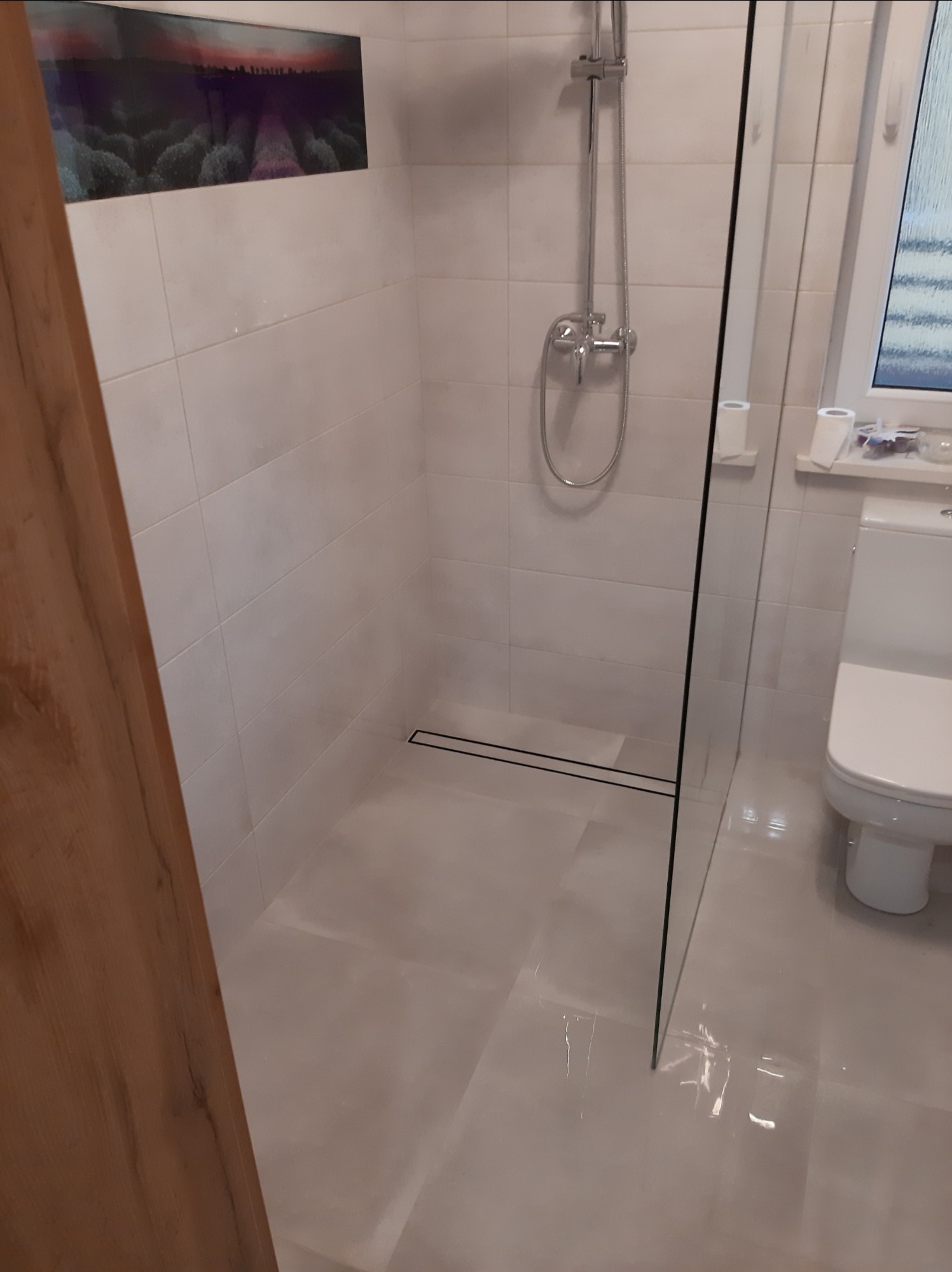 Wykończona kabina prysznicowa z odpływem liniowym i szklaną ścianką, widoczna toaleta i dekoracyjny panel ścienny z motywem krajobrazu.