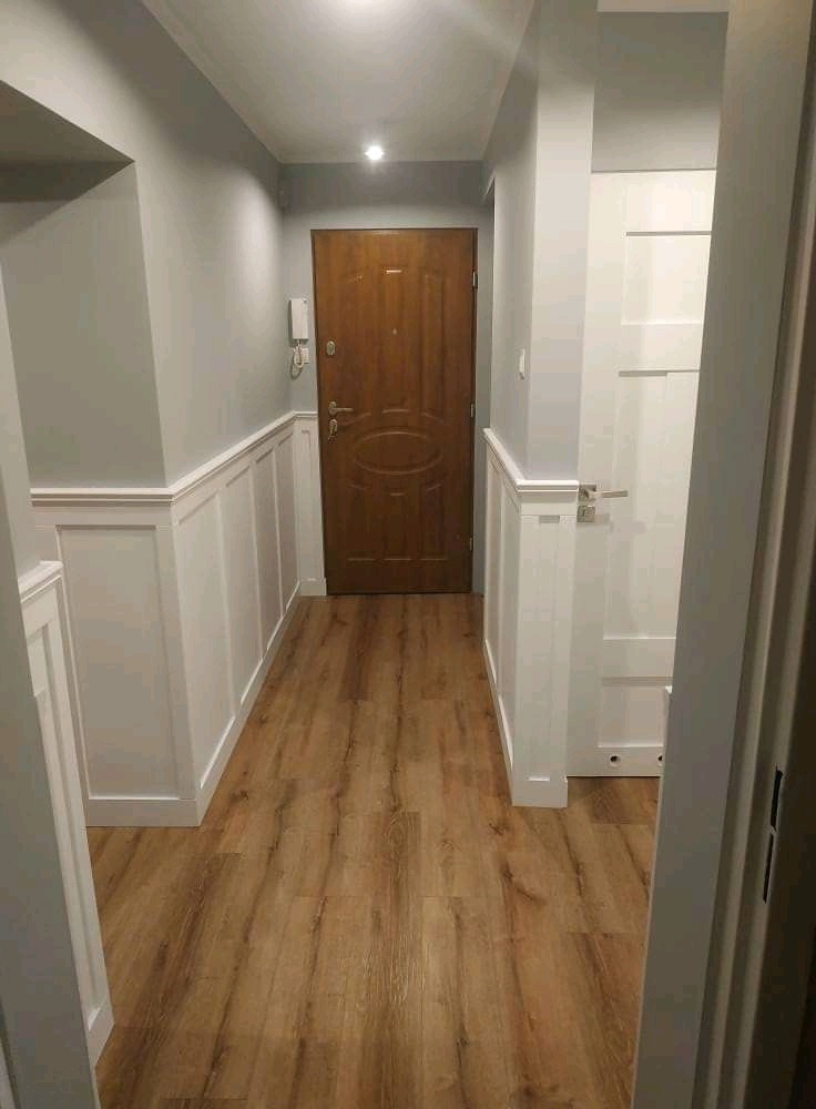 Korytarz po remoncie z drewnianą podłogą imitującą panele, szarą ścianą z białymi panelami dekoracyjnymi i drewnianymi drzwiami wejściowymi.