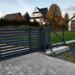 Nowoczesna, przesuwna brama ogrodzeniowa z automatyką, oświetlona pomarańczową lampką, na tle kostki brukowej i domów jednorodzinnych.