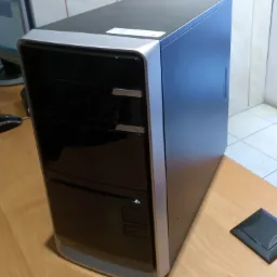 Komputer stacjonarny typu tower w obudowie czarno-niebieskiej z srebrnymi obramowaniami, umieszczony na drewnianym biurku, obok widoczny element biurkowy.