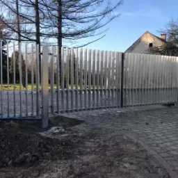 Nowoczesna brama wjazdowa ze stali nierdzewnej z pionowymi szczeblami, widok z zewnątrz na tle domu i drzew bez liści, świeżo ułożona kostka brukowa, ziemia wokół fundamentów.