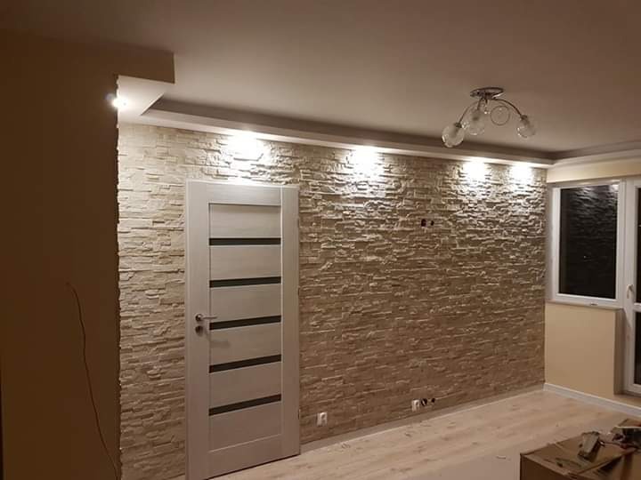 Salon z dekoracyjną ścianą z kamienia elewacyjnego, oświetleniem punktowym i nowoczesnymi drzwiami wewnętrznymi z przeszkleniami, widoczne gniazdka elektryczne i lampa sufitowa.