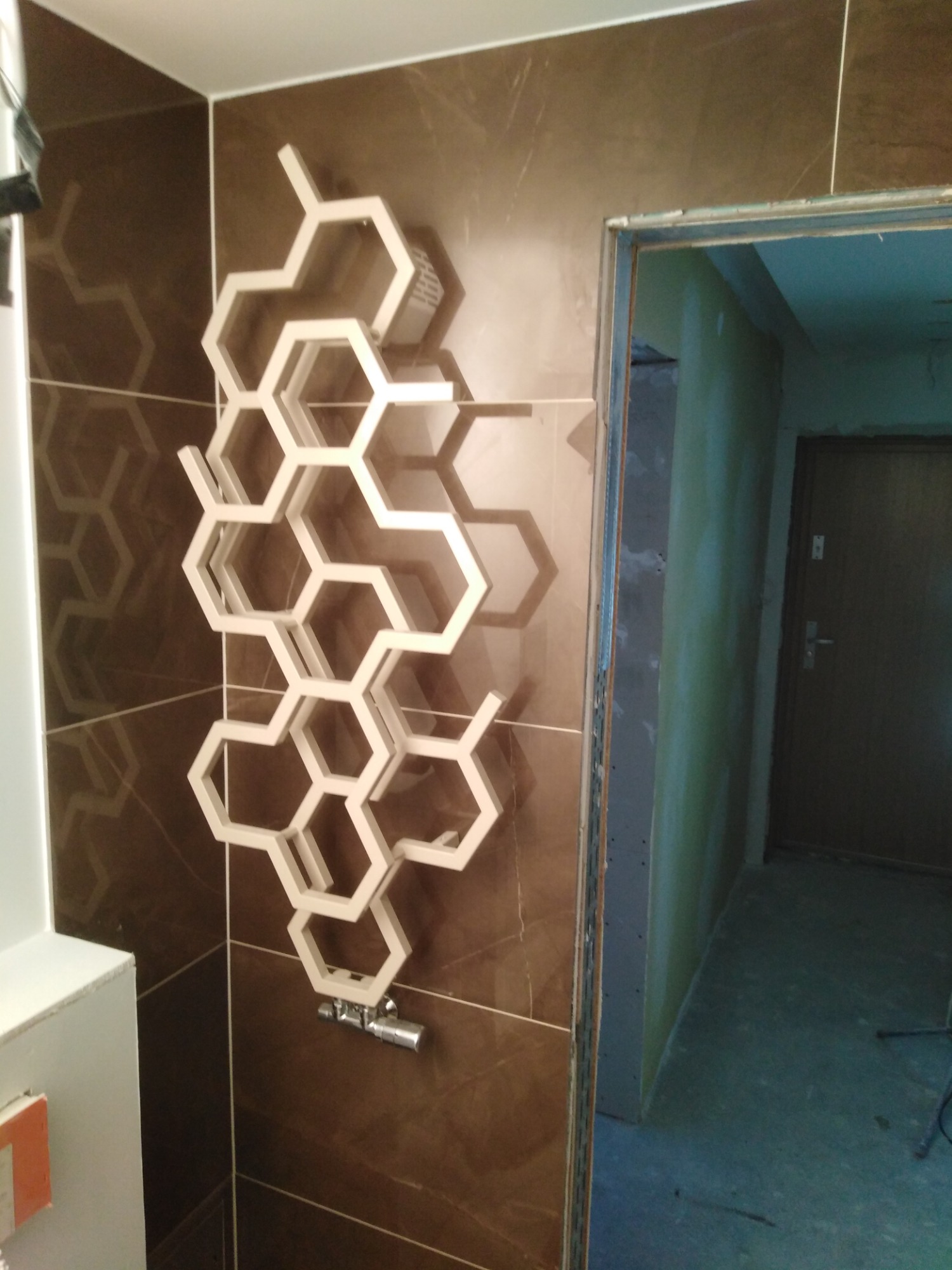 Brązowe płytki imitujące kamień na ścianie łazienki z zamontowanym dekoracyjnym grzejnikiem w kształcie plastra miodu, widok na surową ścianę i drzwi wejściowe.