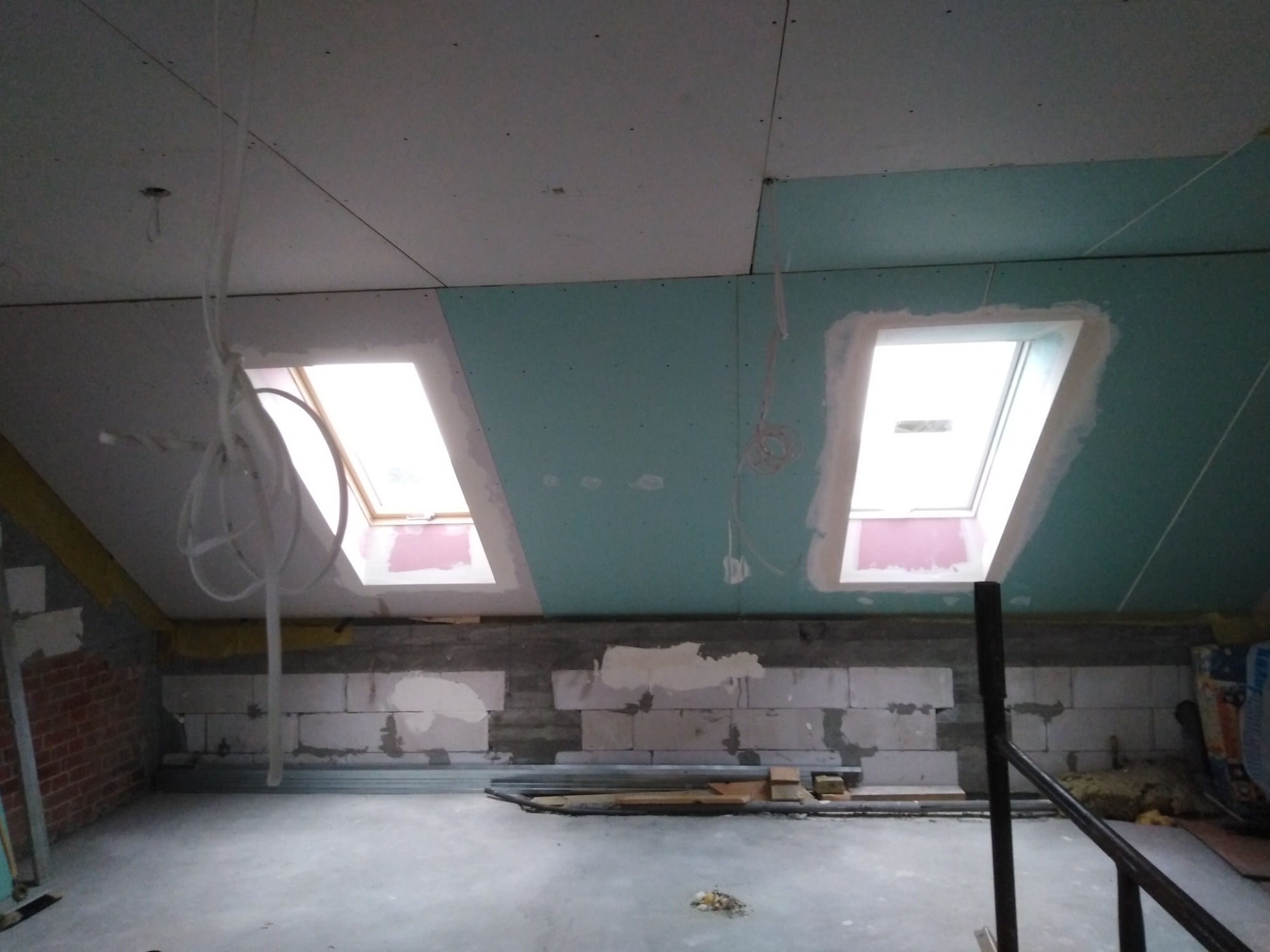 Poddasze w trakcie adaptacji: widoczne okna dachowe, częściowo zamontowana płyta karton-gips na suficie, ściany z bloczków betonowych, elementy konstrukcji i instalacje.