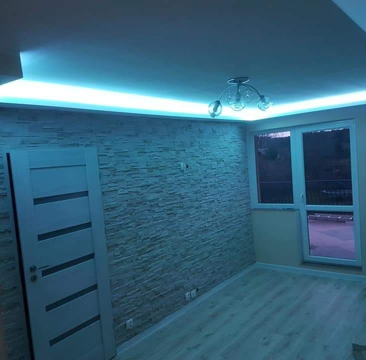 Pokój z kamienną ścianą, oświetleniem LED podsufitowym, panelem podłogowym i drzwiami z dekoracyjnymi frezami.