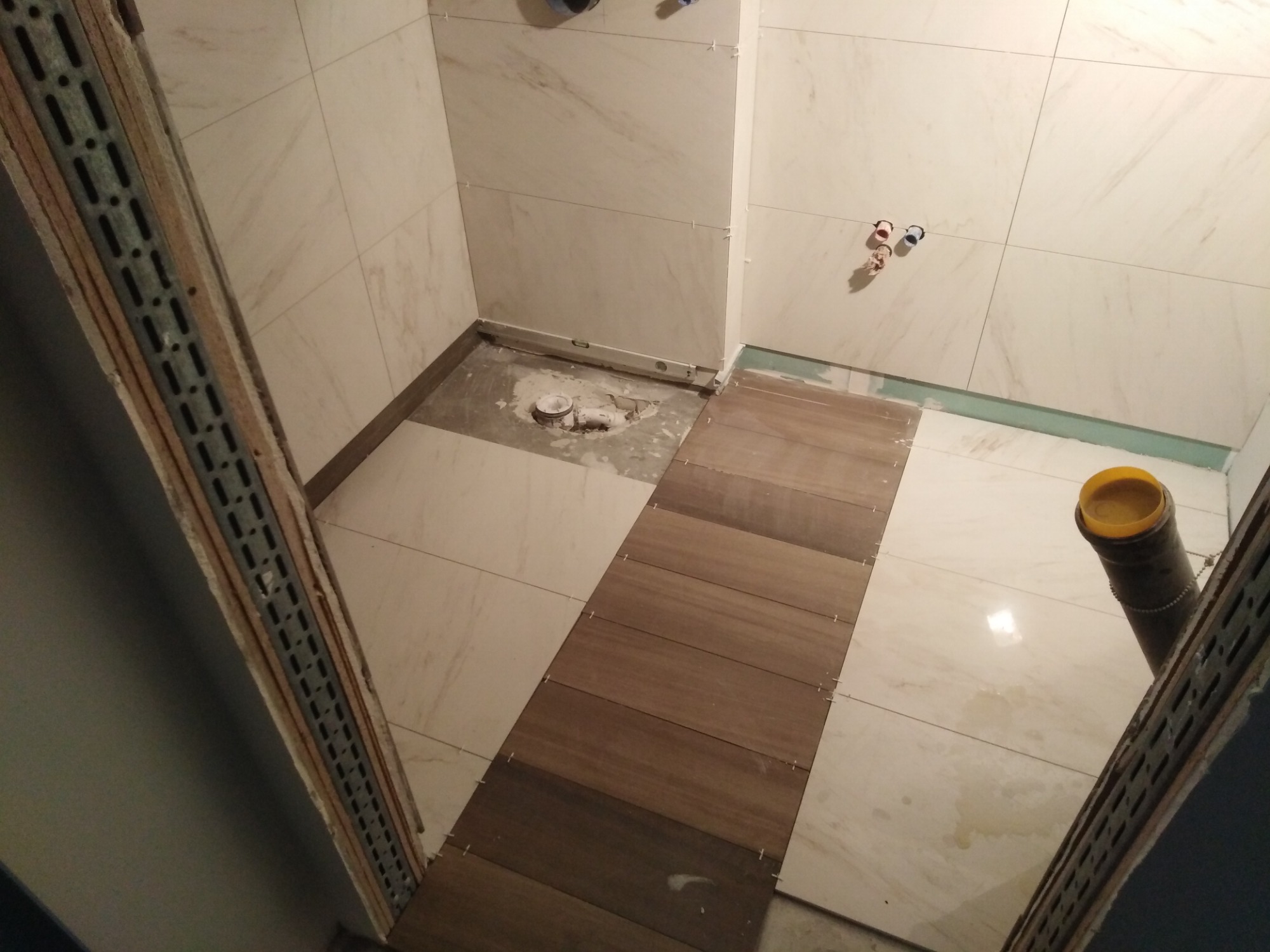 W trakcie układania drewnopodobnych paneli podłogowych w łazience z jasnymi, marmurkowymi płytkami ściennymi. Widoczne krzyżyki dystansowe między panelami, przygotowane podłoże pod prysznic i rury...