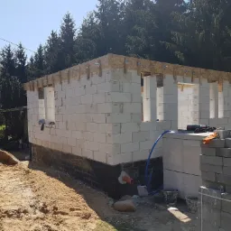 Wznoszenie ścian parterowego domu z bloczków betonu komórkowego, widoczne zbrojenie wieńca i izolacja fundamentów, obok hałda piasku i paleta z bloczkami.