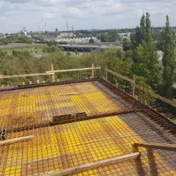 Zbrojenie stropu na żółtym szalunku, widok z góry na konstrukcję z drewnianymi barierkami, w tle panorama miasta z kominami i zielenią.