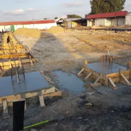 Zalane betonem szalunki fundamentowe z wystającymi zbrojeniami na placu budowy, w tle różowa betoniarka i hałdy piasku.