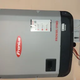 Szary inwerter Fronius Symo zamontowany na ścianie obok dwóch skrzynek elektrycznych z widocznymi przewodami. Na inwerterze widoczny wyświetlacz z wartością 529 oraz naklejki informacyjne.