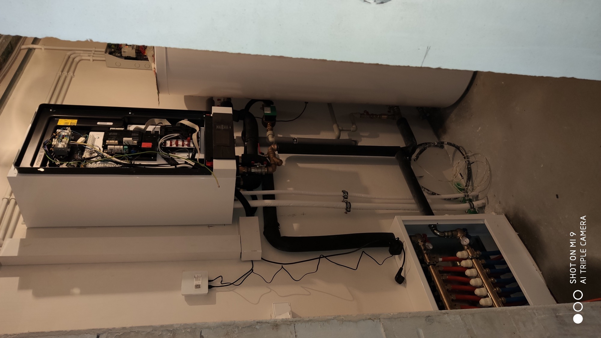 Rozbudowana instalacja grzewcza z otwartym panelem sterującym, widoczne liczne rury miedziane i plastikowe, zbiornik ciepłej wody oraz rozdzielacz z zaworami, całość w surowym pomieszczeniu...