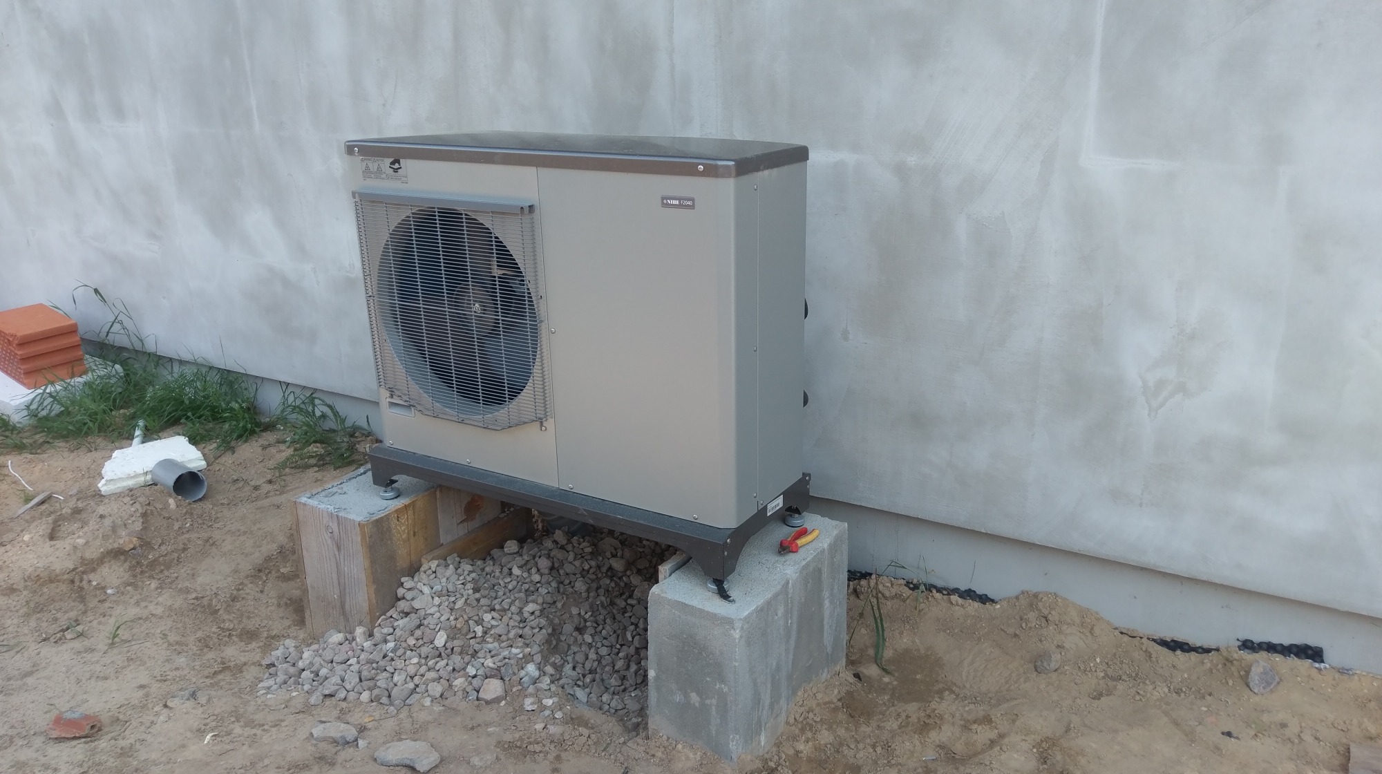 Zewnętrzna jednostka pompy ciepła Nibe 12040, zamontowana na betonowych bloczkach, z widocznymi narzędziami (szczypce) obok, na tle surowej ściany budynku.