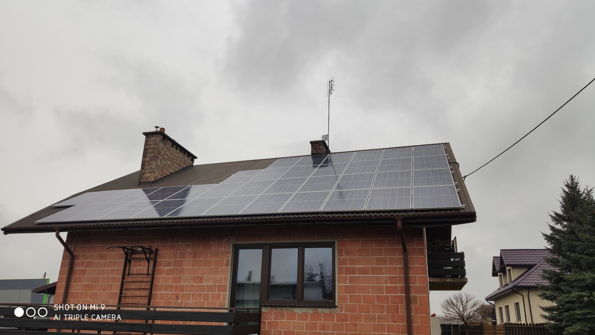 Brązowy dach domu z cegły z zamontowanymi panelami słonecznymi w pochmurny dzień, widoczny komin i antena.