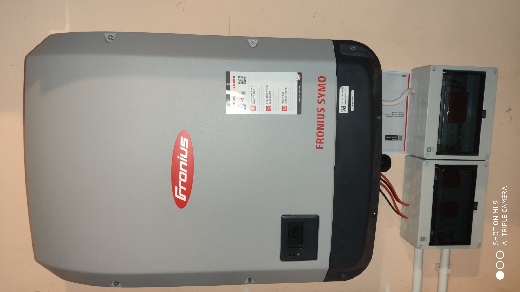 Szary inwerter Fronius Symo zamontowany na ścianie obok dwóch skrzynek elektrycznych z widocznymi przewodami. Na inwerterze widoczny wyświetlacz z wartością 529 oraz naklejki informacyjne.