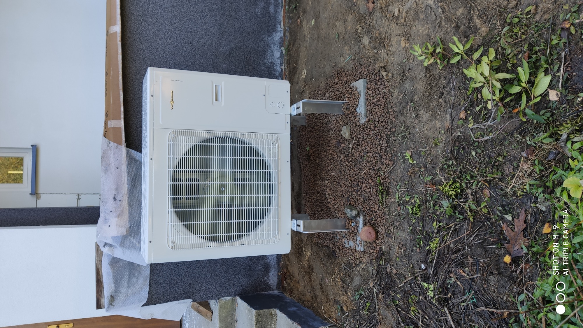 Biała jednostka zewnętrzna pompy ciepła Viessmann zamontowana na metalowych wspornikach na gruncie wysypanym drobnymi kamykami, widoczne elementy budynku w tle.