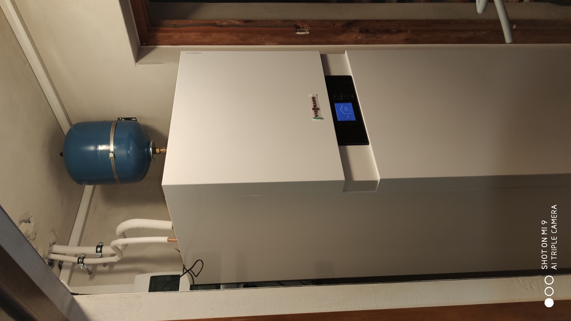 Biała pompa ciepła marki Viessmann z niebieskim zbiornikiem wyrównawczym zamontowana w pomieszczeniu z drewnianym elementem wykończeniowym. Widoczne rury w izolacji.