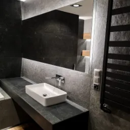 Nowoczesna łazienka z umywalką nablatową, baterią chromowaną i grzejnikiem drabinkowym na tle ściany wyłożonej szarymi płytkami imitującymi kamień. W lustrze odbija się fragment drewnianych półek...