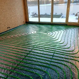 Zielone rurki ogrzewania podłogowego rozłożone na czarnej macie w pomieszczeniu z oknem i ścianami wykończonymi płytami OSB, widok na zimowy krajobraz za oknem.