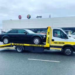Czarny samochód osobowy załadowany na platformę pojazdu pomocy drogowej z napisem 'POMOC DROGOWA' i numerem telefonu, na parkingu przed salonem samochodowym wielu marek.