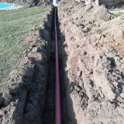 Wykopany rów z ułożoną rurą kanalizacyjną z tworzywa sztucznego, na powierzchni ziemi widoczna koparka i hałdy ziemi.