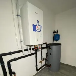 Biały piec grzewczy Panasonic z naklejką w kształcie kciuka w górę, podłączony do czarnych rur instalacyjnych i szarego zbiornika akumulacyjnego w pomieszczeniu z szarą podłogą.