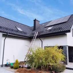 Nowoczesny dom jednorodzinny z ciemną dachówką i panelami słonecznymi na dachu, drabina oparta o rynnę, widoczne narzędzia i wiadro sugerujące prace instalacyjne.