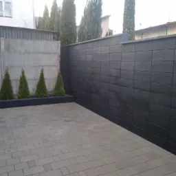 Ułożona szara kostka brukowa na podwórku z widocznym betonowym i czarnym murem oraz rzędem małych tujek, z elementami zabudowy w tle.
