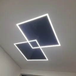 Geometryczna oprawa sufitowa z dwoma kwadratami LED, z których jeden nachodzi na drugi, oświetlając wnętrze. Widoczny fragment okna i ściany.