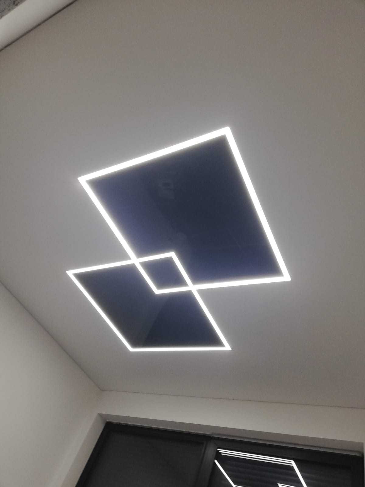 Geometryczna oprawa sufitowa z dwoma kwadratami LED, z których jeden nachodzi na drugi, oświetlając wnętrze. Widoczny fragment okna i ściany.
