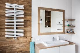 Nowoczesna łazienka z drewnopodobnymi płytkami, białym grzejnikiem drabinkowym, prostokątną umywalką nablatową i lustrem w drewnianej ramie. Nad umywalką zwisają dwie szklane lampy na czarnych...