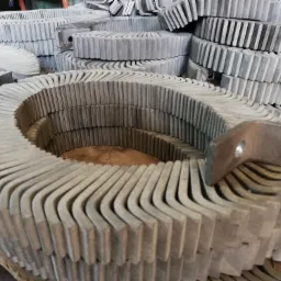 Stos ułożonych, szarych, metalowych elementów giętych w kształt litery L, z widocznym spawem i otworem na śrubę, tworzących okrągłe segmenty.