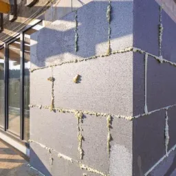 Szare bloki styropianowe z pianką montażową, tworzące ocieplenie ściany zewnętrznej budynku w trakcie budowy, widoczne drewniane elementy konstrukcji dachu i okno.