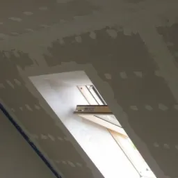 Montaż płyty gipsowo-kartonowej na poddaszu, widoczne łączenia płyt szpachlowane wokół okna dachowego, niebieska listwa narożna na krawędzi ściany.