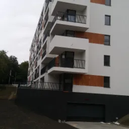 Nowoczesny budynek wielorodzinny w Siemianowicach Śląskich z białymi elewacjami, balkonami z czarnymi barierkami i elementami dekoracyjnymi imitującymi drewno. Widoczny parking z kostki brukowej.