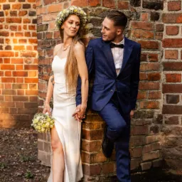 Para młoda, on w granatowym garniturze w kratę, ona w prostej sukni z rozcięciem i wiankiem, trzymają się za ręce, opierając się o ceglany mur.