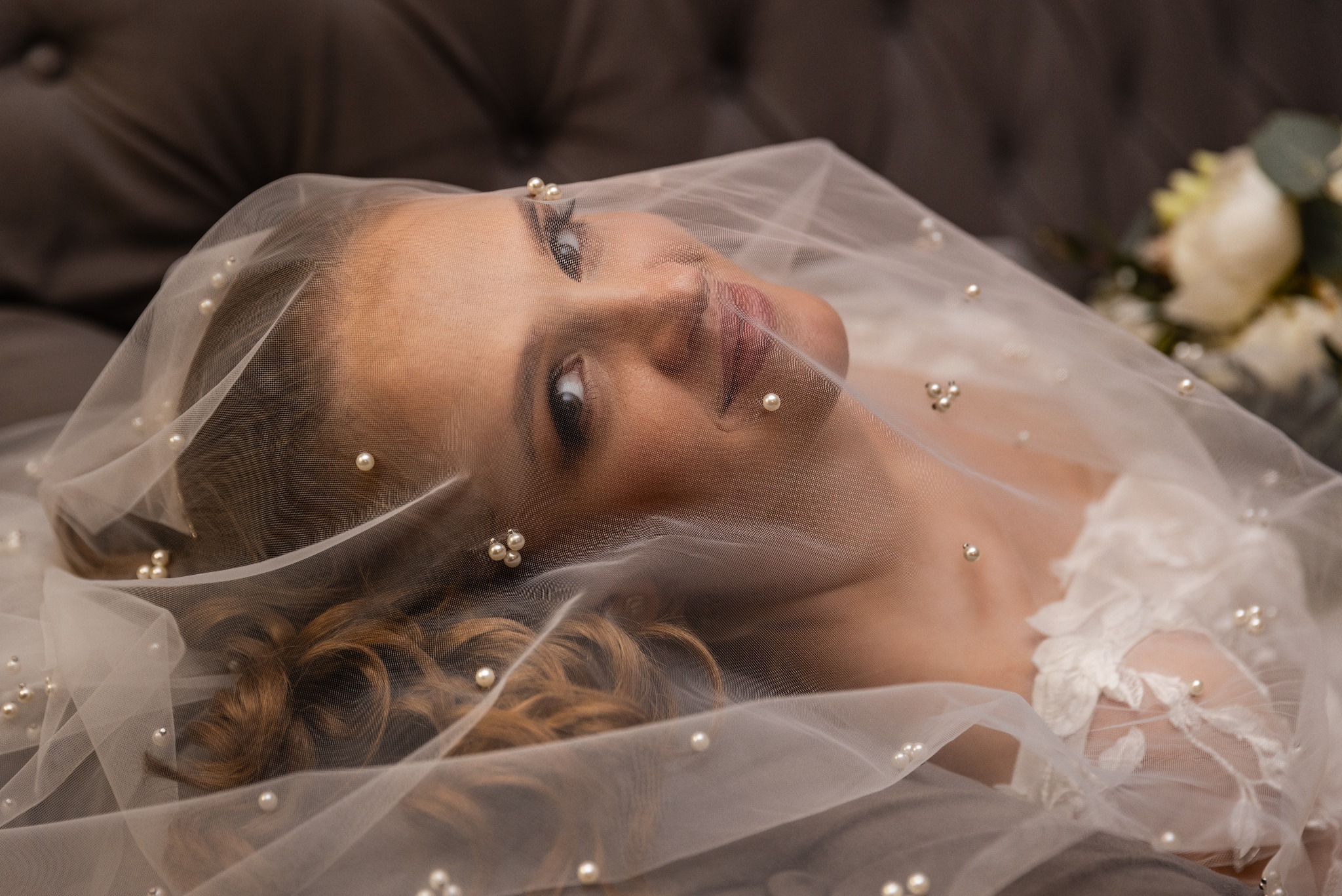 Panna młoda leżąca na szarym, pikowanym fotelu, z welonem zdobionym perłami zakrywającym twarz, w tle bukiet ślubny.