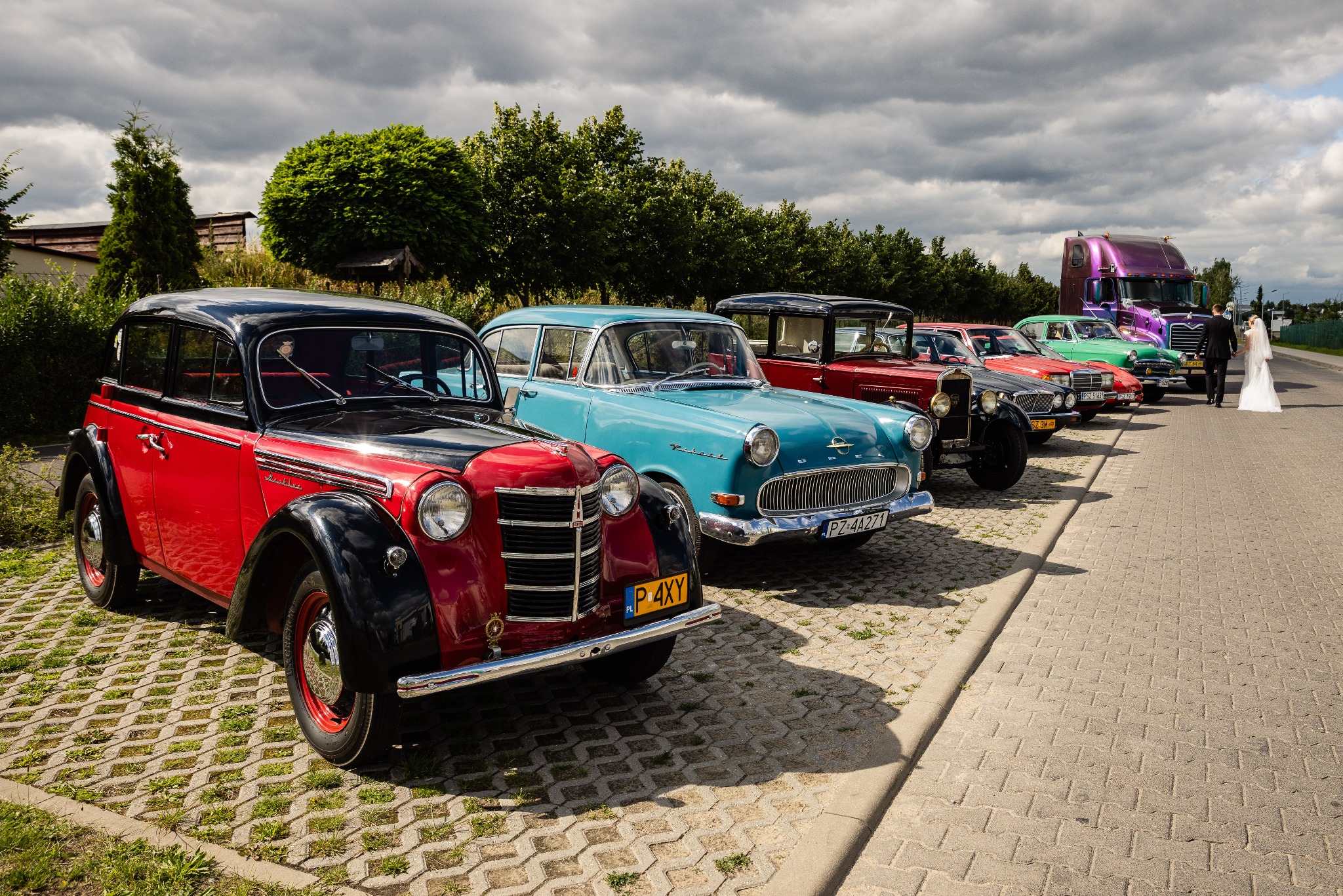 Para młoda w oddali idzie w kierunku rzędu zabytkowych, kolorowych samochodów, w tym czerwonego Moskwicza i turkusowego Opla, zaparkowanych na brukowanej drodze, z fioletową ciężarówką w tle.