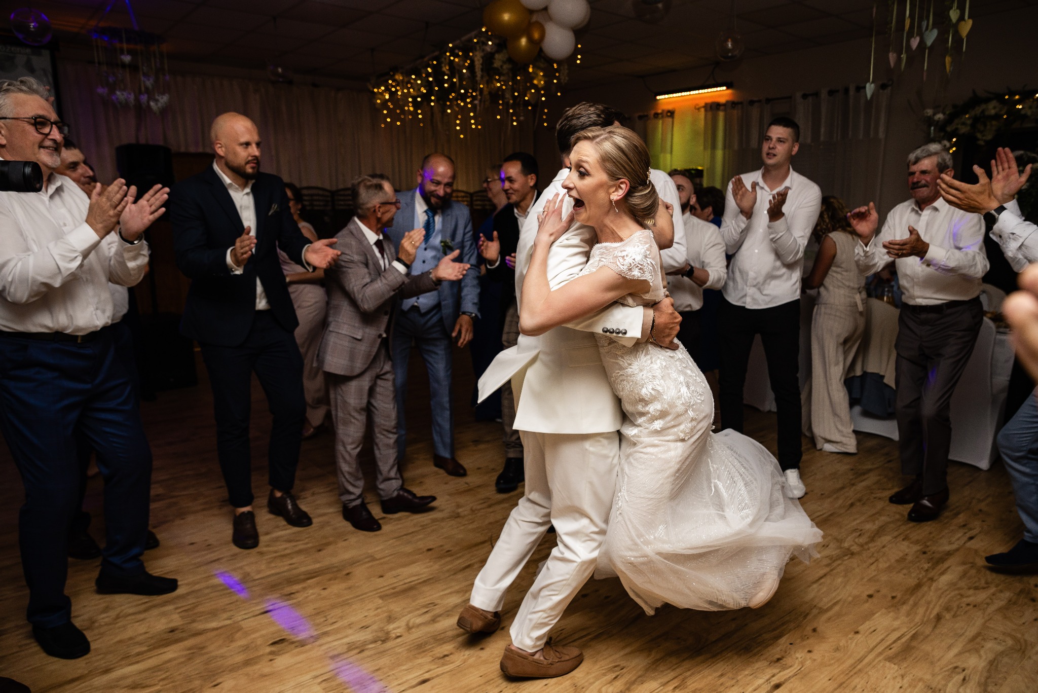 Para młoda w objęciach podczas pierwszego tańca, otoczona rozentuzjazmowanym tłumem gości weselnych na parkiecie z drewnianą podłogą, oświetlonym dekoracyjnymi lampkami i balonami.