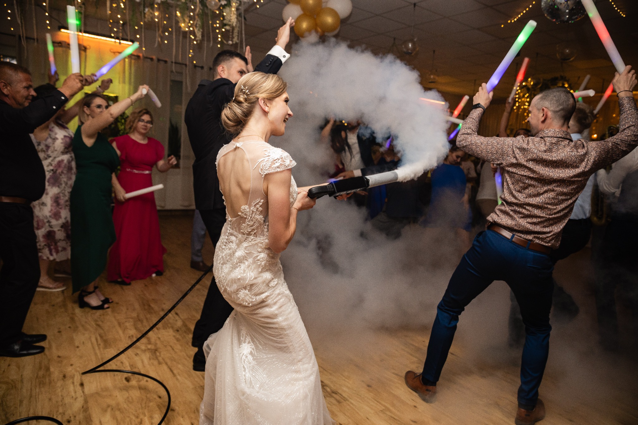 Panna młoda w koronkowej sukni, uśmiechnięta, trzyma wyrzutnię dymu na parkiecie podczas wesela, w tle goście z ledowymi pałkami.
