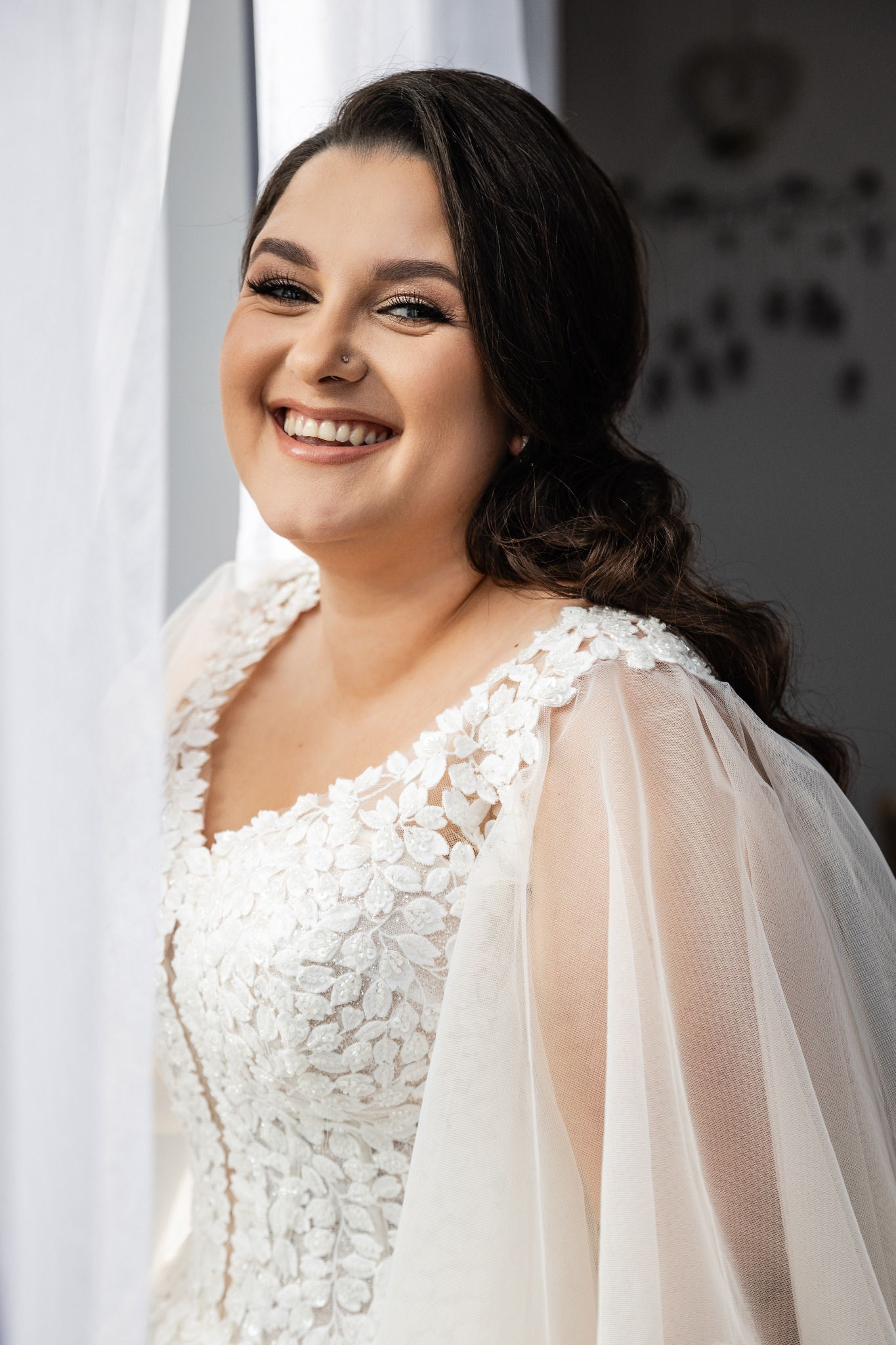Uśmiechnięta panna młoda w koronkowej sukni z tiulowymi rękawami, z kolczykiem i piercingiem w nosie, pozuje przy oknie z białymi zasłonami.