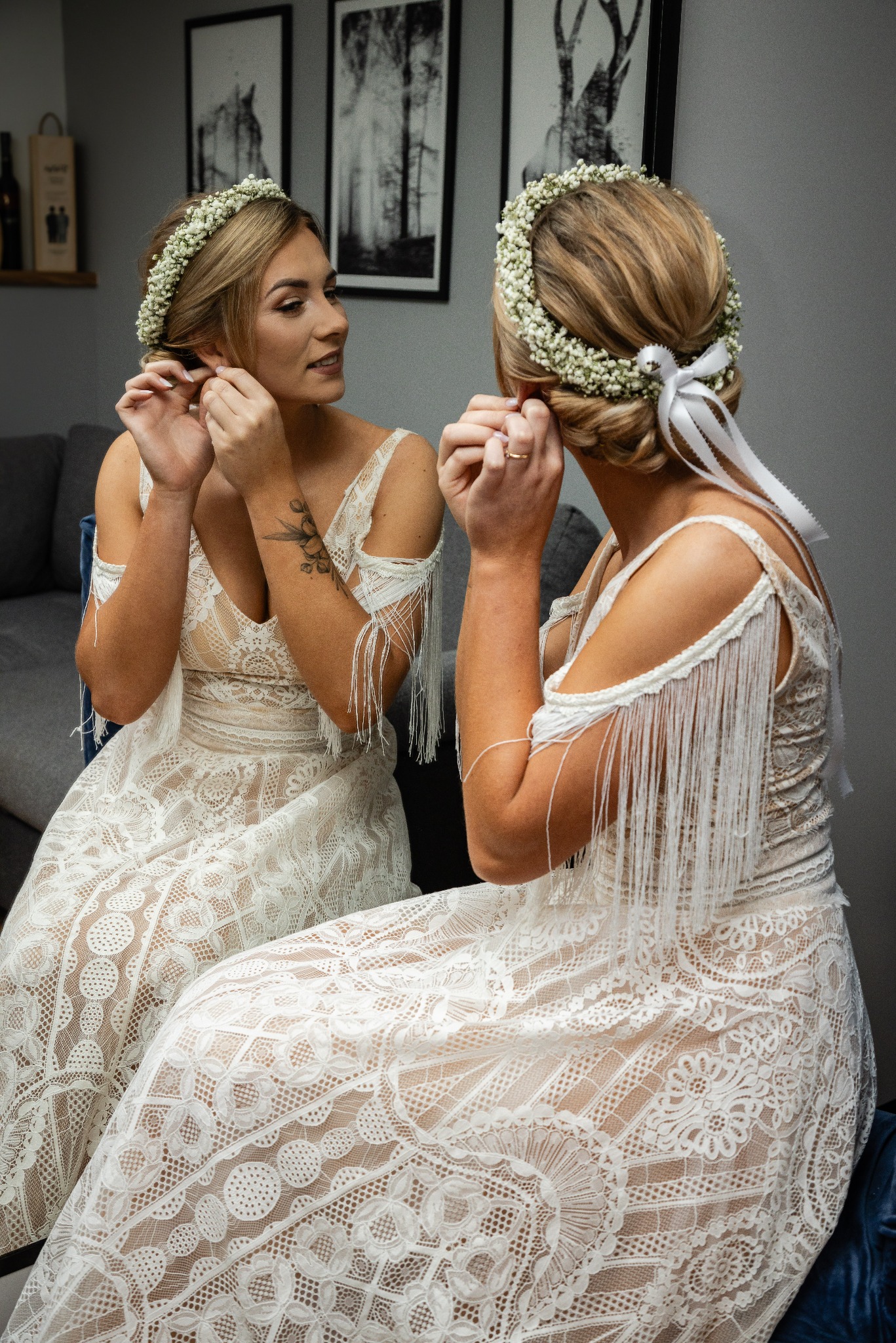 Panna młoda w koronkowej sukni z frędzlami, wiankiem z białych kwiatów i wstążką, przegląda się w lustrze, zakładając kolczyk. Widoczne tatuaże na ramieniu i pierścionek na palcu.