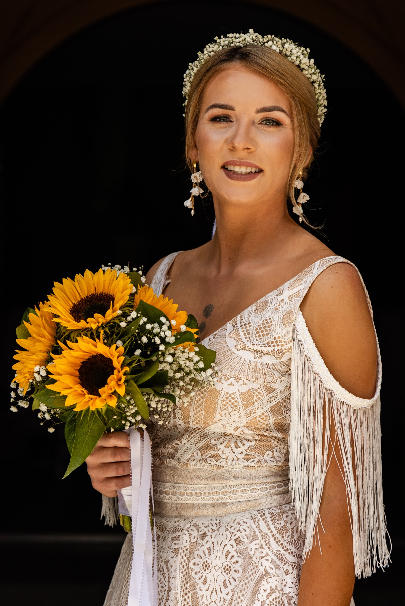 Uśmiechnięta panna młoda w koronkowej sukni z frędzlami na ramionach, trzyma bukiet słoneczników i gipsówki, ozdobiona wiankiem z gipsówki i kolczykami z perłami, na czarnym tle.