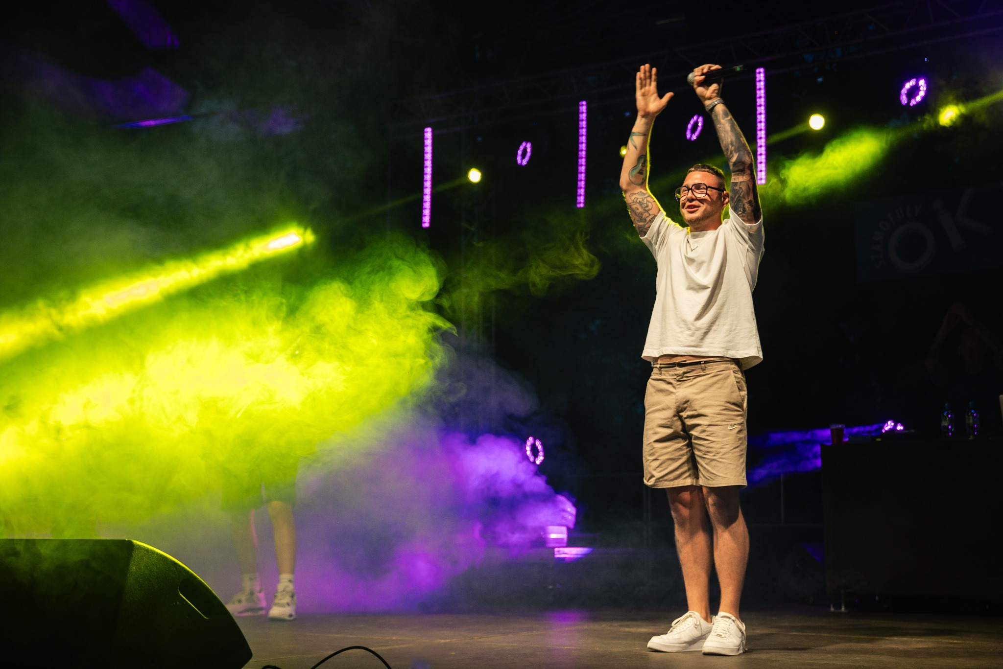 Mężczyzna z tatuażami na scenie, w okularach, z mikrofonem w ręku, unosi ręce w górę, w tle efekty świetlne i dym.