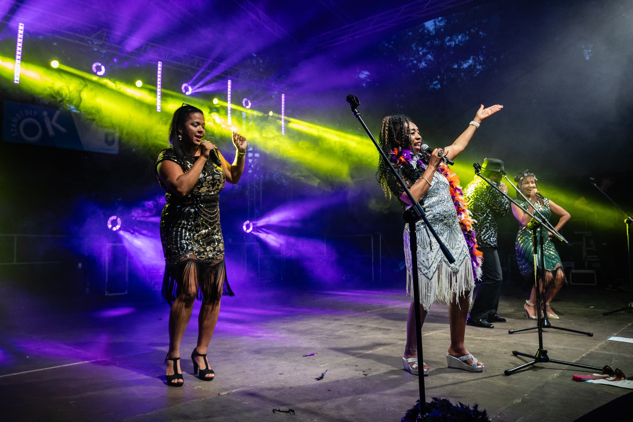 Zespół muzyczny w stylu lat 20. występuje na scenie podczas wydarzenia, oświetlony fioletowymi i żółtymi światłami, wokalistki w cekinowych sukienkach z frędzlami i mikrofonami.